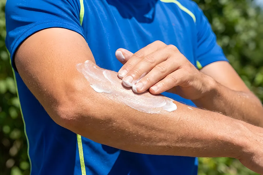 man applying white protective sunscreen lotion onto his arm while wearing a bright blue shirt outdoors in summer