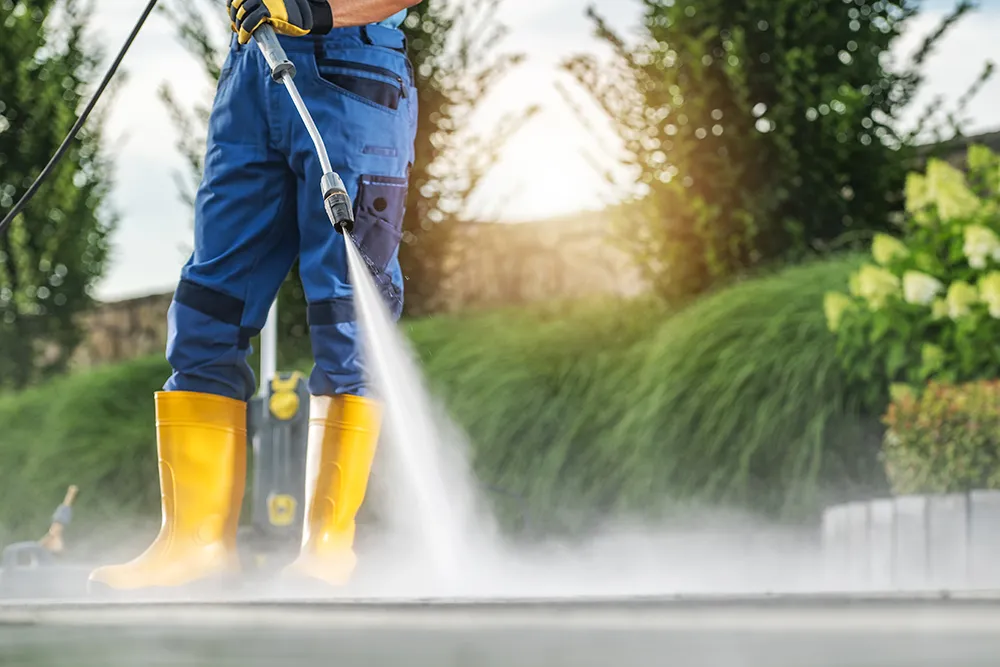 Man Using a Pressure Washer to Clean Outdoor Patio During Bright Sunny Day in a Landscaped Backyard